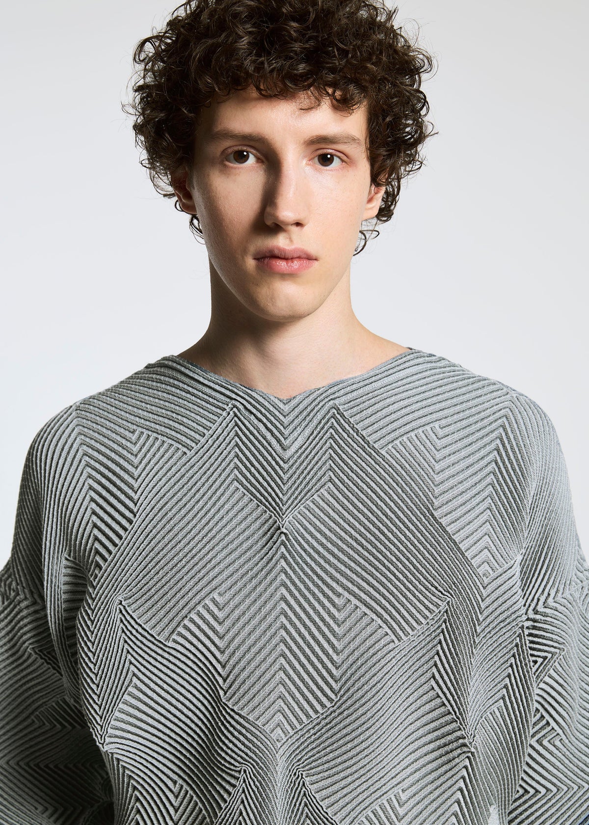 A young person with short, curly brown hair wears the TYPE-O 010 textured light gray sweater by A-POC ABLE ISSEY MIYAKE, featuring a subtle houndstooth pattern. They look at the camera against a plain, light background. view 3 of 6