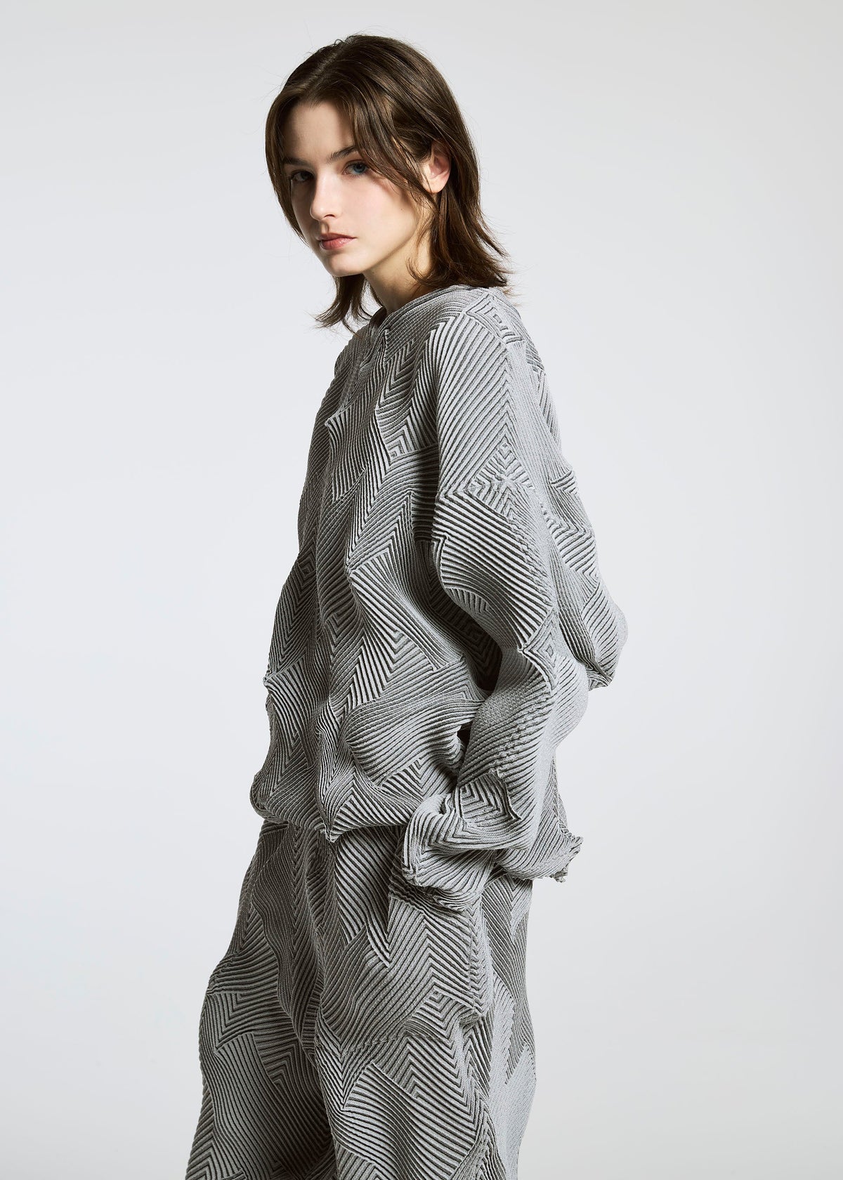 A person with shoulder-length brown hair models the TYPE-O 010 by A-POC ABLE ISSEY MIYAKE, a loose-fitting gray and white houndstooth outfit, standing sideways and looking at the camera against a plain light background. view 4 of 6