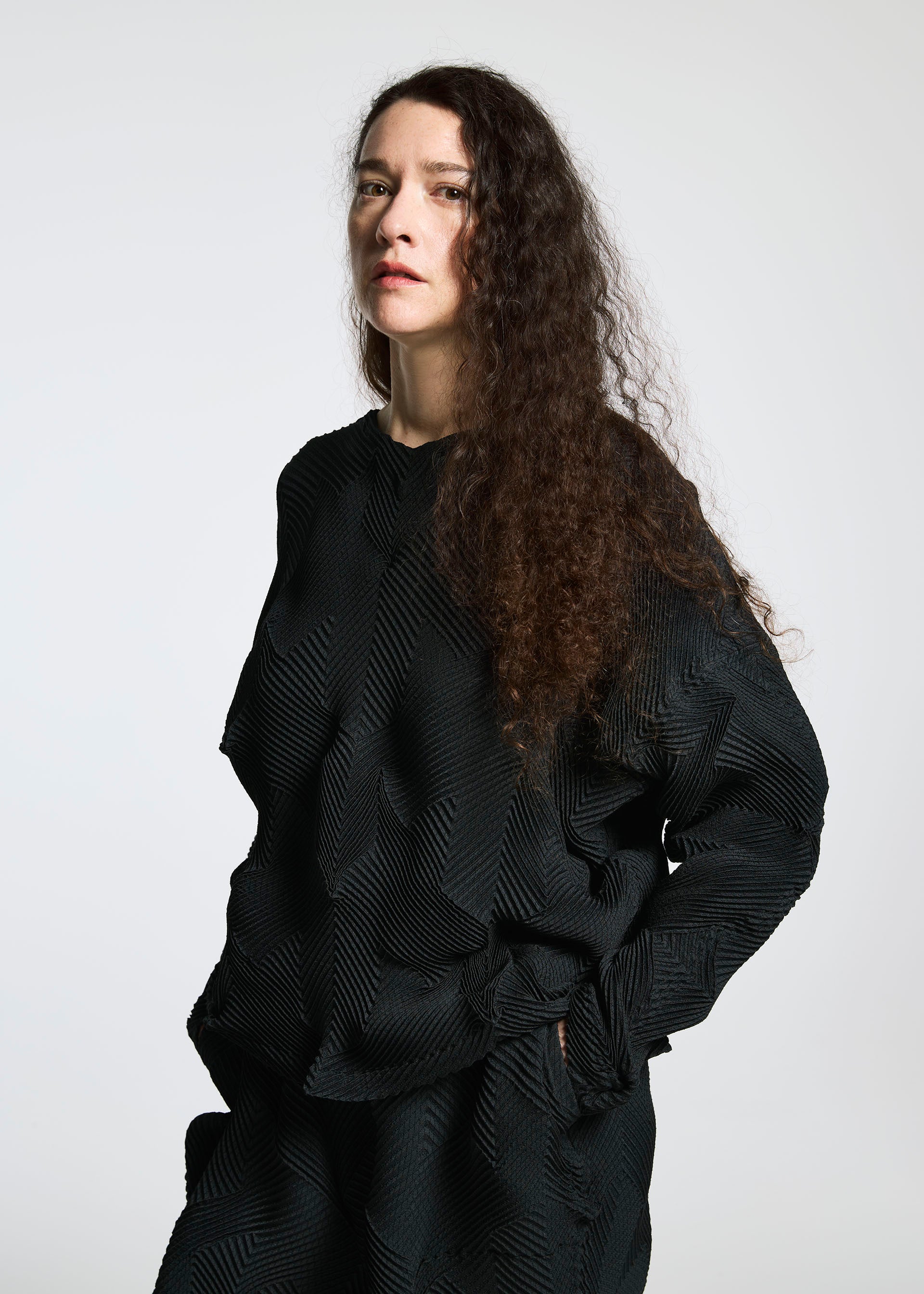 Zoomed view A person with long, curly brown hair wears the TYPE-O 010 pleated black top by A-POC ABLE ISSEY MIYAKE, made from recycled polyester, standing against a light background and looking slightly past the camera with a neutral expression.
