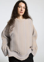 A person with long, wavy brown hair wears the TYPE-W 010 sweater by A-POC ABLE ISSEY MIYAKE in beige with a knit-like texture and black pants, standing against a plain light background and looking directly at the camera.