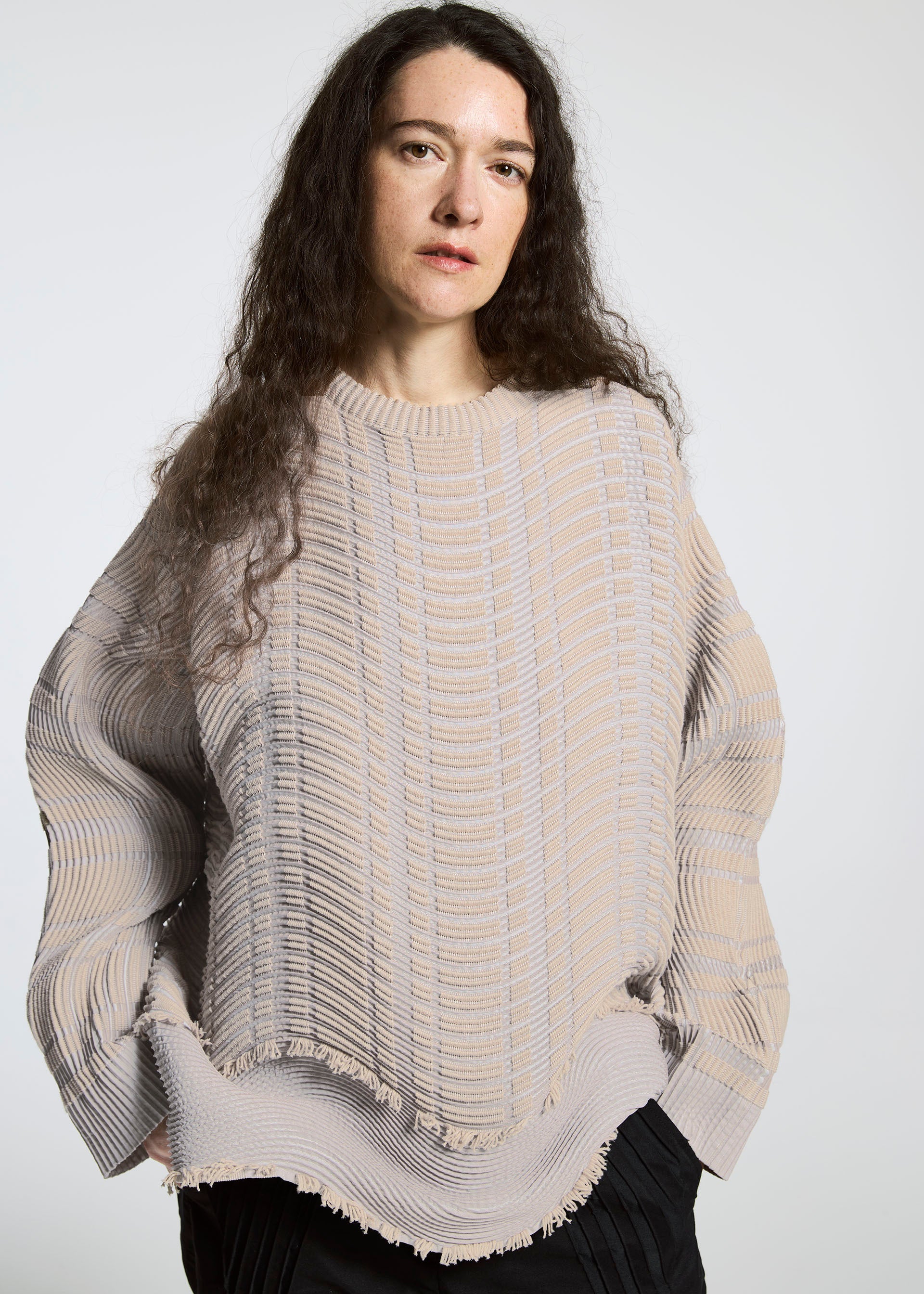 Zoomed view A person with long, wavy brown hair wears the TYPE-W 010 sweater by A-POC ABLE ISSEY MIYAKE in beige with a knit-like texture and black pants, standing against a plain light background and looking directly at the camera.