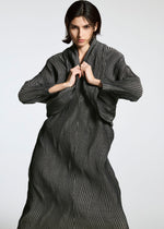 A person with short dark hair wears the TYPE-XI Spiber project dress by A-POC ABLE ISSEY MIYAKE, a textured, biodegradable dark gray piece with wavy vertical stripes, holding the collar against a plain light background.