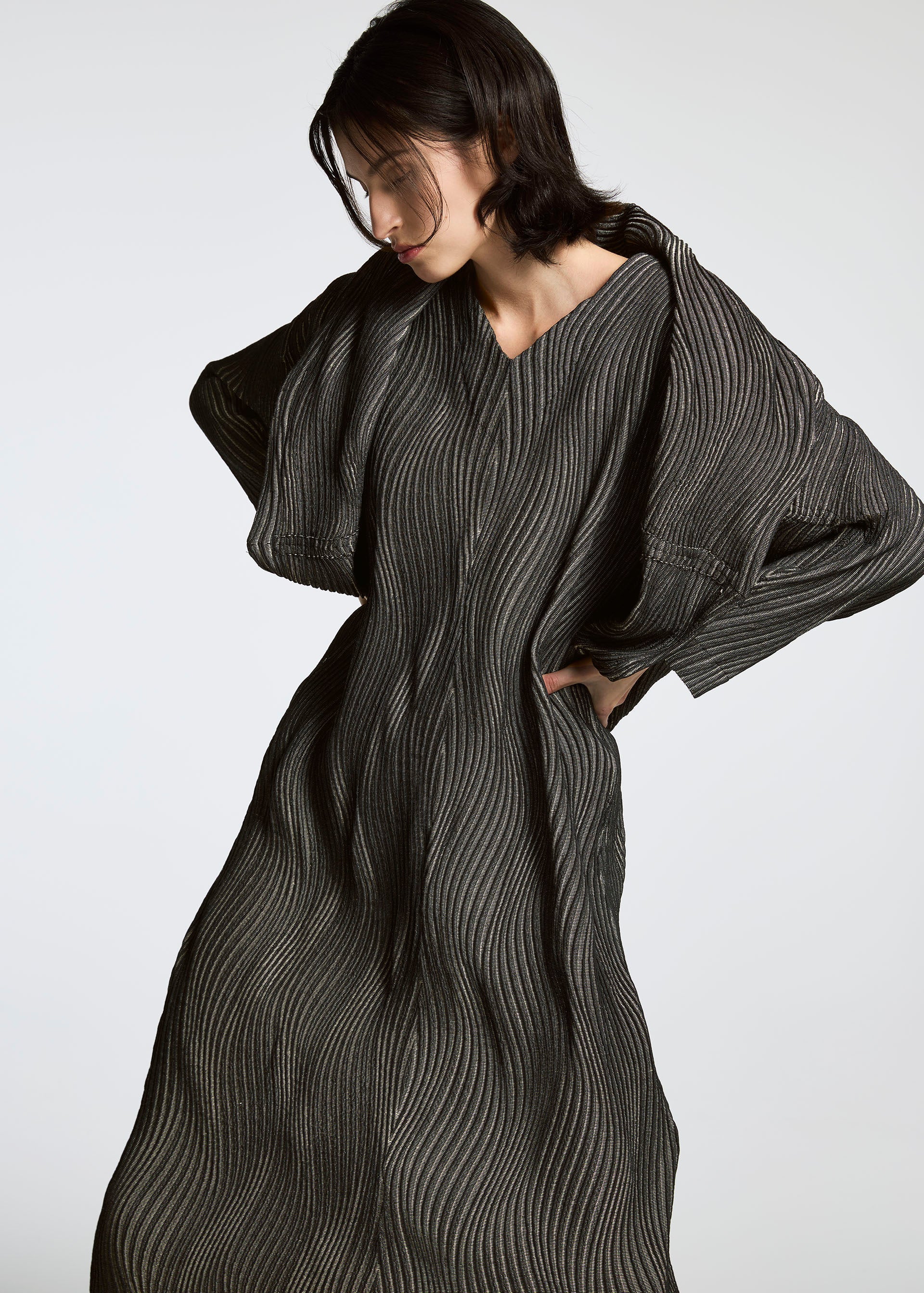Zoomed view A person with short dark hair wears a long, flowing TYPE-XI Spiber project dress by A-POC ABLE ISSEY MIYAKE, featuring wavy vertical stripes. They stand against a plain light background, looking down with bent arms and hands behind their back.
