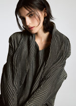 A person with dark, chin-length hair poses in the TYPE-XI Spiber project by A-POC ABLE ISSEY MIYAKE, a pleated Brewed Protein™ fiber garment in gray and silver, set against a plain light background and looking thoughtfully at the camera.