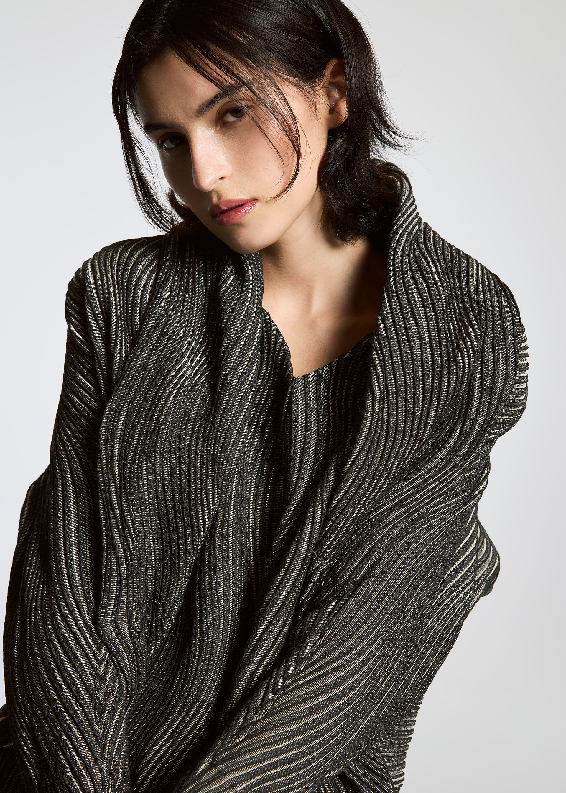 Zoomed view A person with dark, chin-length hair poses in the TYPE-XI Spiber project by A-POC ABLE ISSEY MIYAKE, a pleated Brewed Protein™ fiber garment in gray and silver, set against a plain light background and looking thoughtfully at the camera.