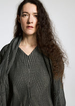 A woman with long, curly brown hair wears a textured, wavy-patterned top and scarf made from Brewed Protein™ fiber by A-POC ABLE ISSEY MIYAKE's TYPE-XI Spiber project, standing against a plain light background with a neutral expression.