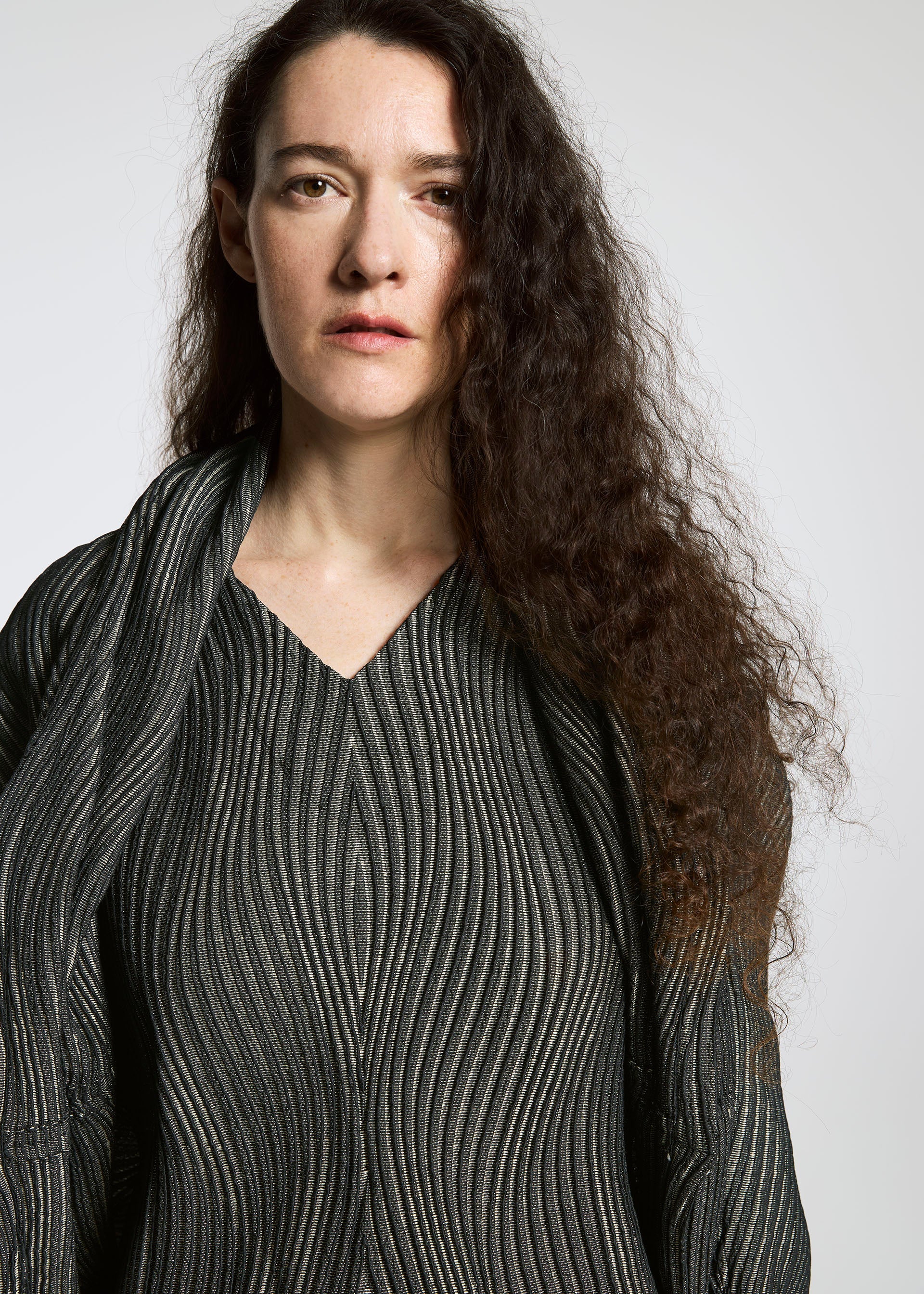 Zoomed view A woman with long, curly brown hair wears a textured, wavy-patterned top and scarf made from Brewed Protein™ fiber by A-POC ABLE ISSEY MIYAKE's TYPE-XI Spiber project, standing against a plain light background with a neutral expression.