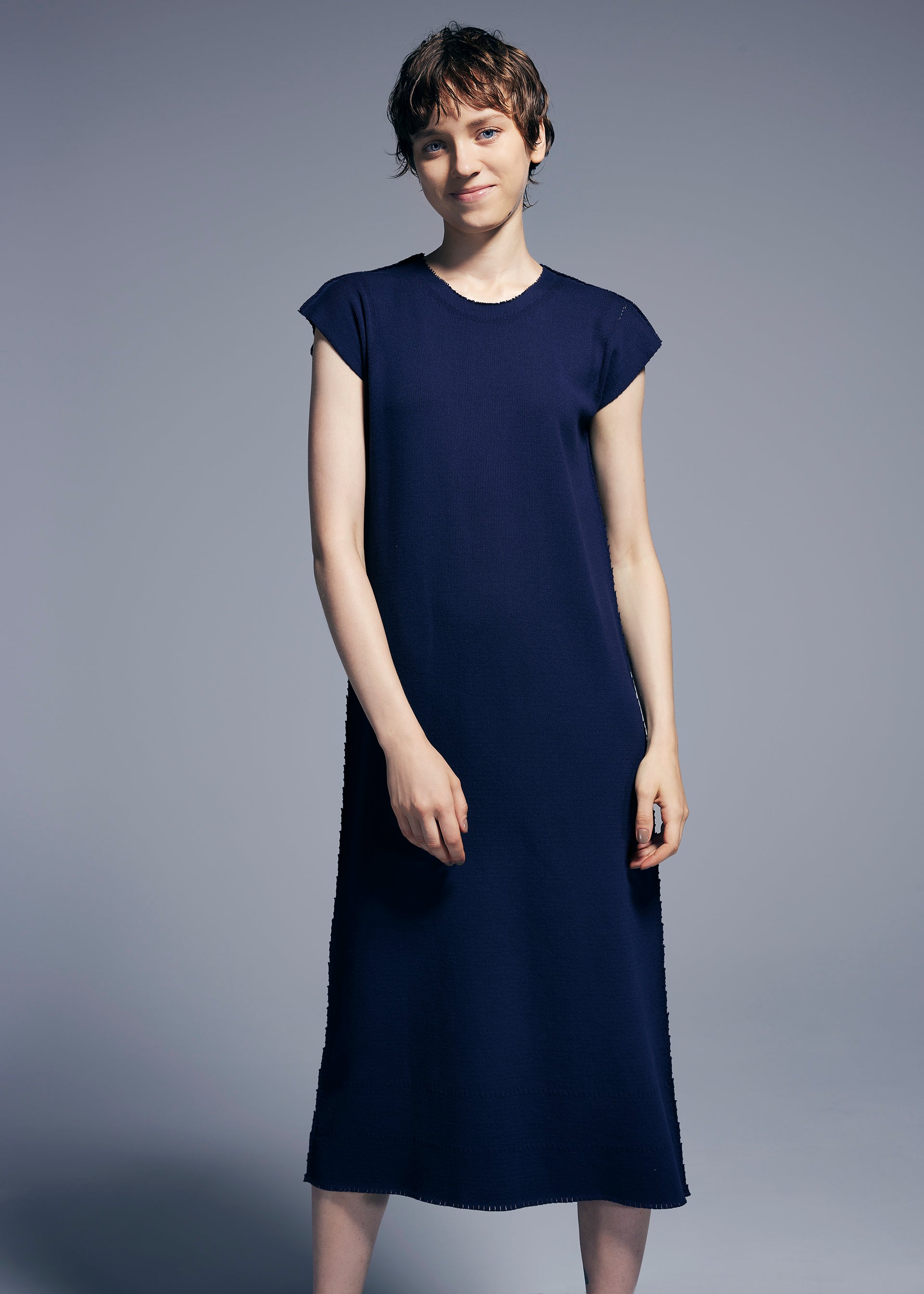 Zoomed view A person with short brown hair wears the TYPE-A BASICS dress by A-POC ABLE ISSEY MIYAKE—a simple, round-neck French sleeve design in navy blue—standing against a plain gray background and smiling softly at the camera.