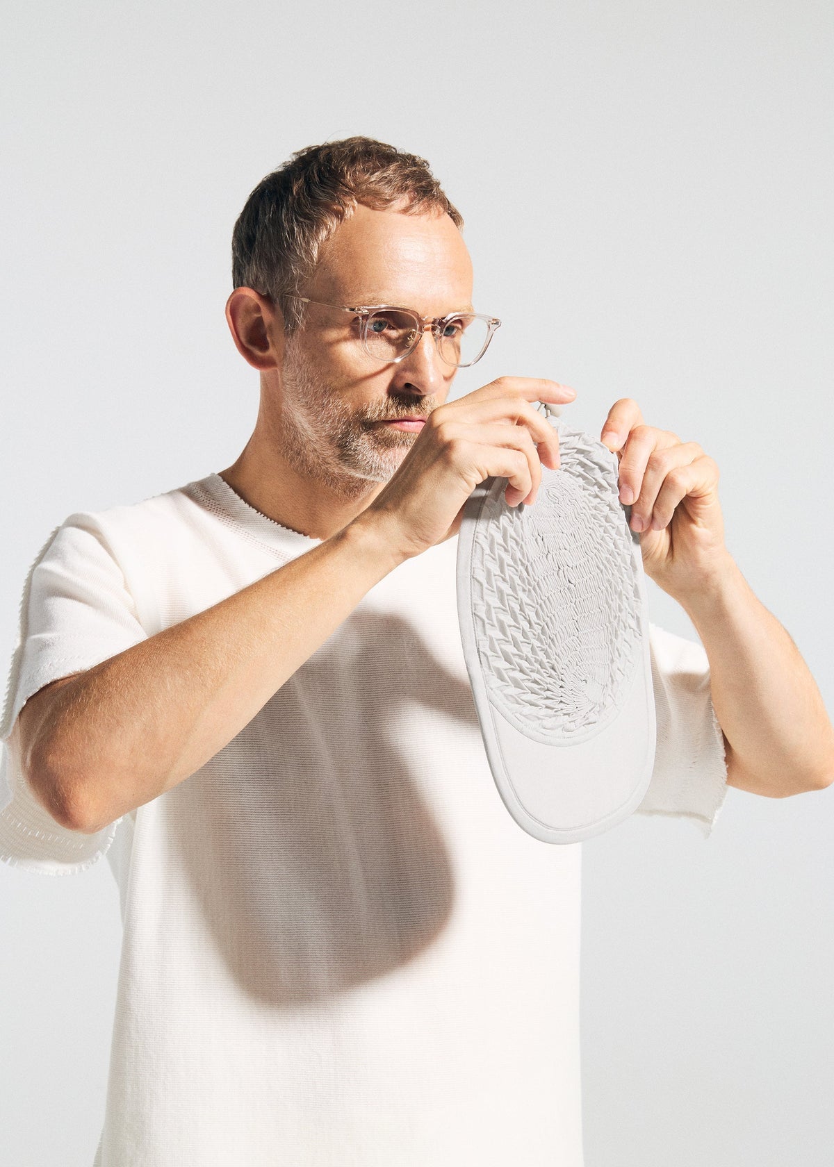 A man in glasses and a white shirt holds up the TYPE-O CAP by A-POC ABLE ISSEY MIYAKE, displaying its intricate geometric pleats made with the Steam Stretch technique, set against a plain light background. view 12 of 15