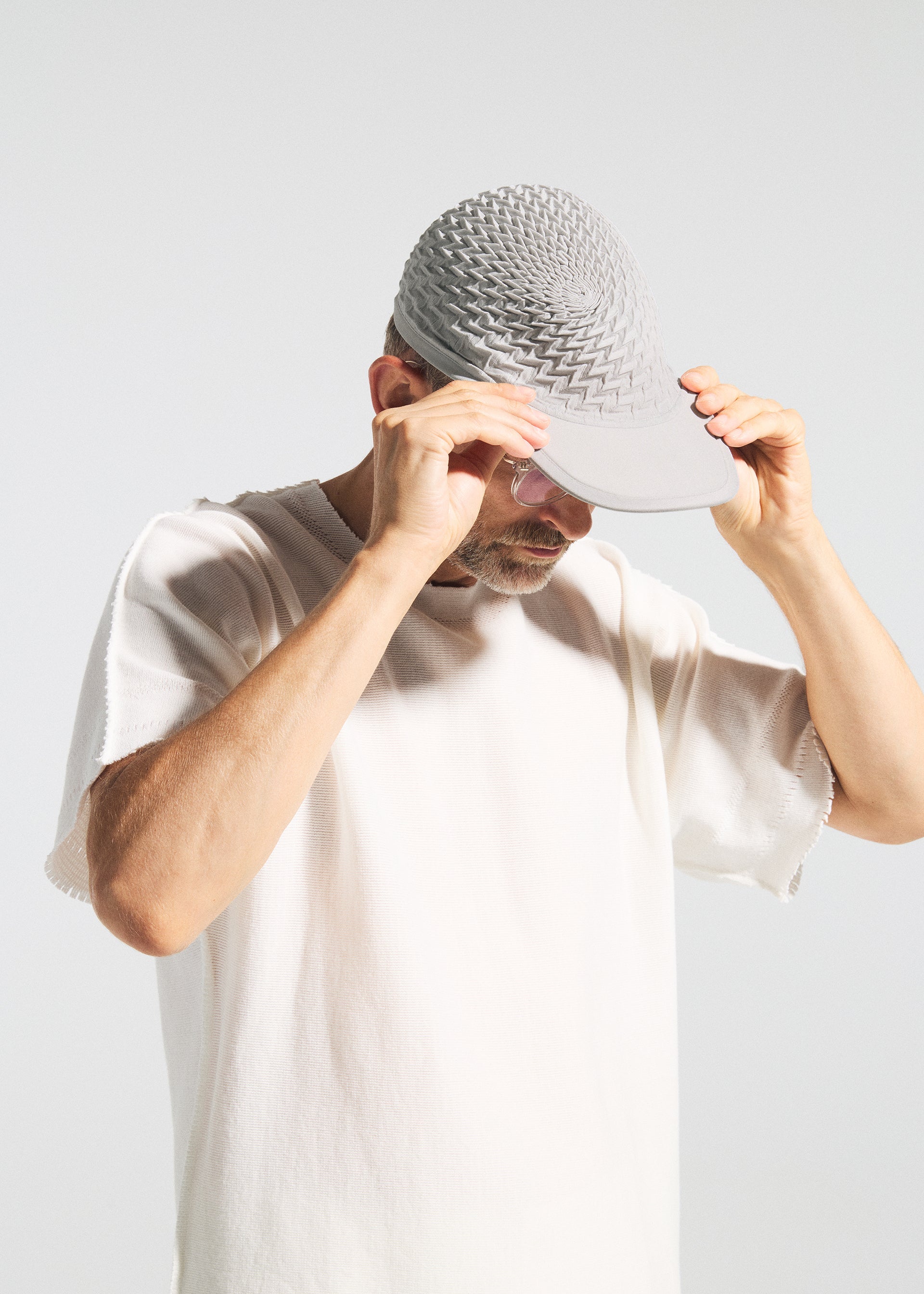 Zoomed view A person in a white shirt adjusts the TYPE-O CAP by A-POC ABLE ISSEY MIYAKE, featuring a light gray, geometric pleated 3D design made with the Steam Stretch technique. The background is plain and light-colored.