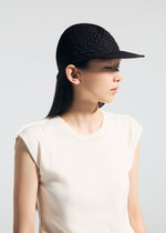 A woman wearing the TYPE-O CAP by A-POC ABLE ISSEY MIYAKE, featuring geometric pleats made with the Steam Stretch technique, and a sleeveless white top looks to her left against a plain light background.