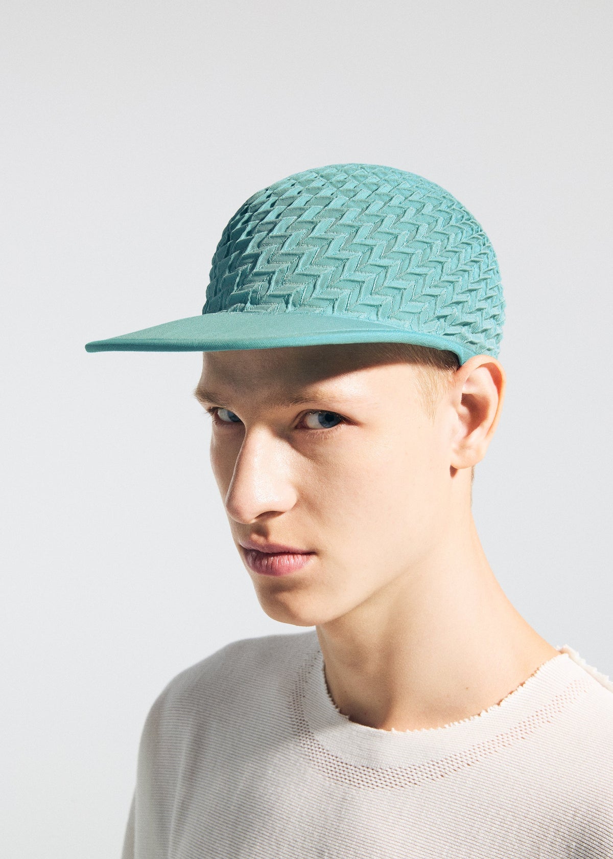 A young person wears the TYPE-O CAP by A-POC ABLE ISSEY MIYAKE, featuring a light teal geometric pleated design with zigzag pattern, paired with a light distressed sweater against a plain background. view 15 of 15