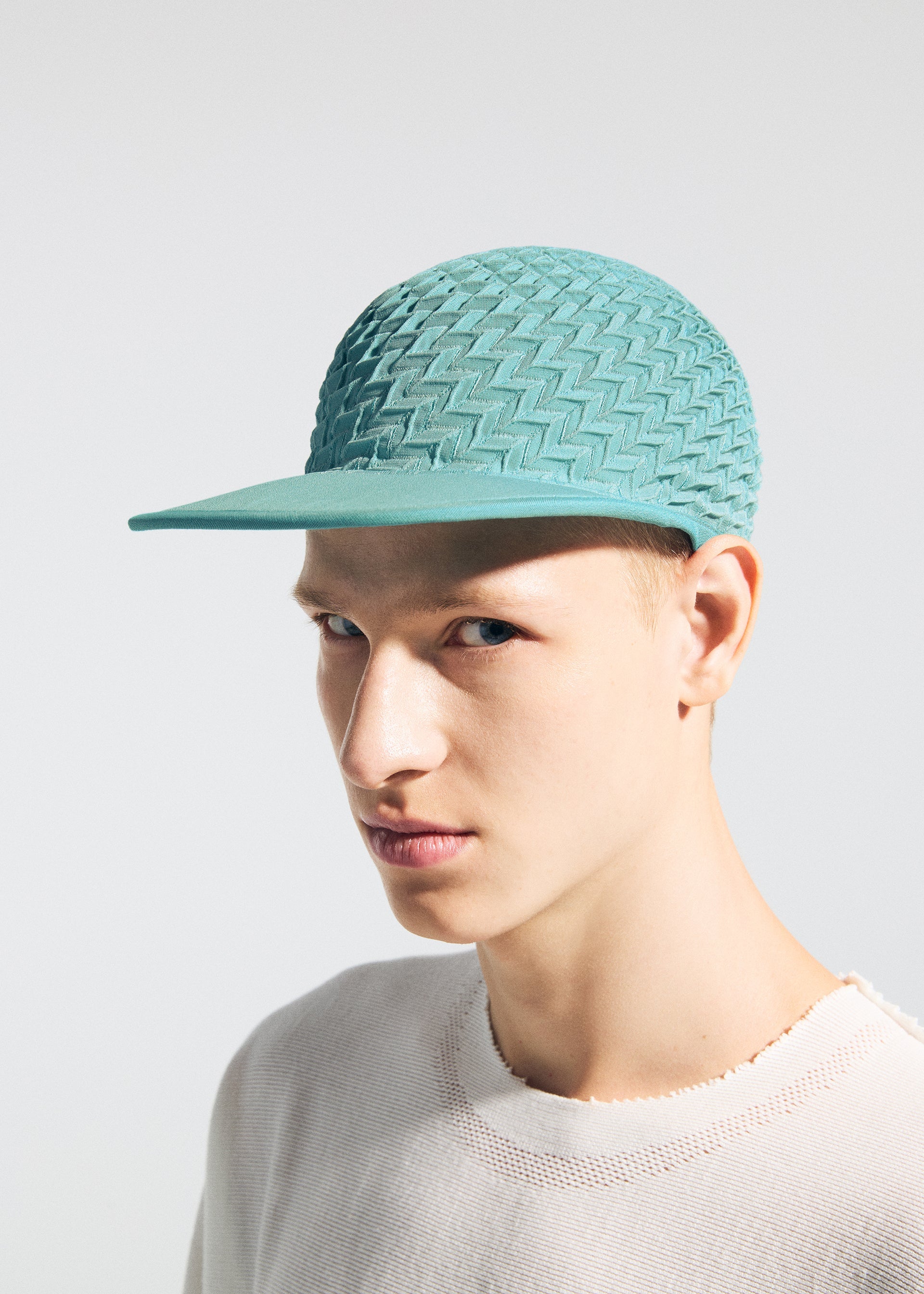 Zoomed view A young person wears the TYPE-O CAP by A-POC ABLE ISSEY MIYAKE, featuring a light teal geometric pleated design with zigzag pattern, paired with a light distressed sweater against a plain background.