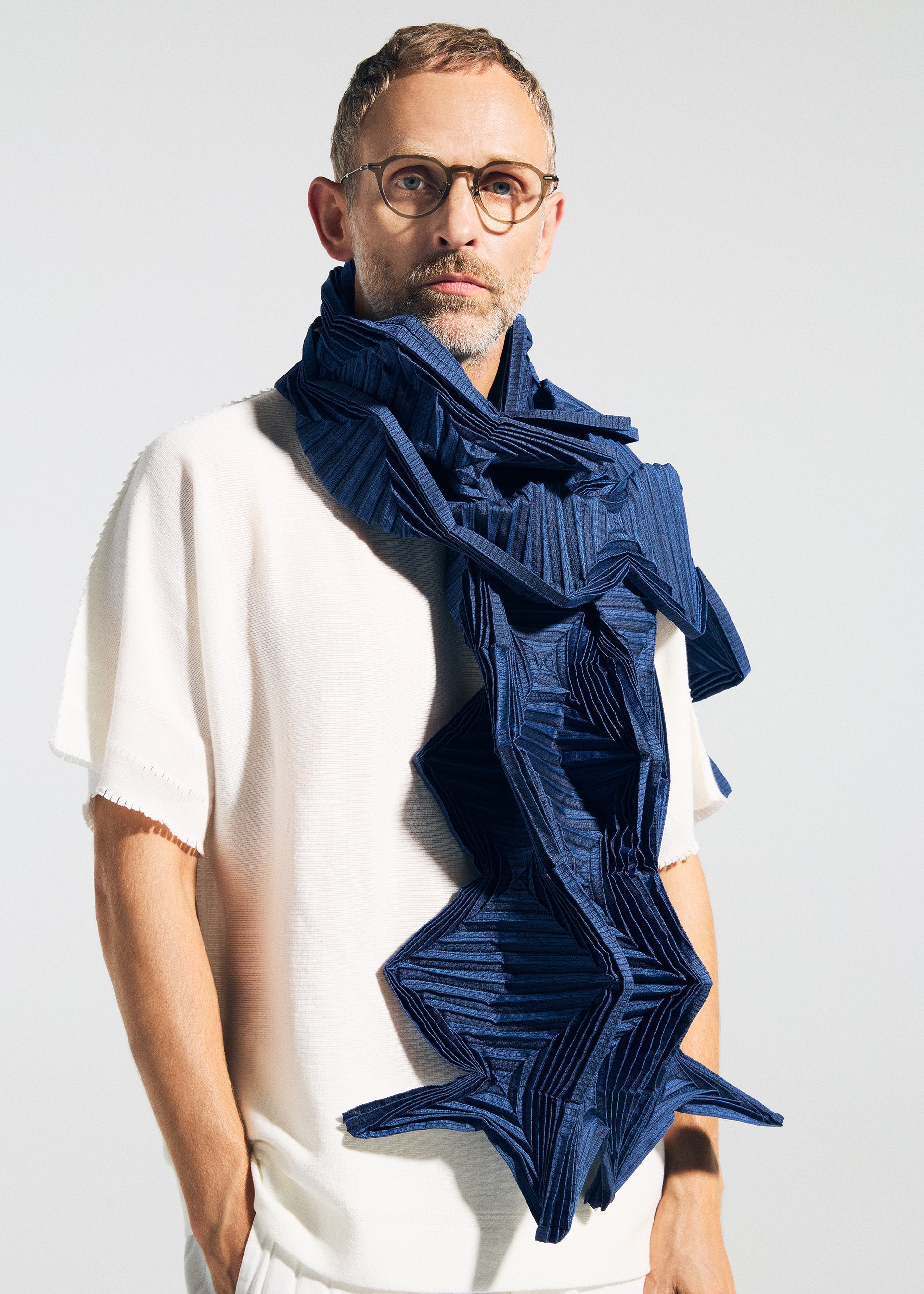Zoomed view A man with short hair, glasses, and a light beard wears a white short-sleeved shirt and the TYPE-O STOLE 002 by A-POC ABLE ISSEY MIYAKE—a large blue pleated stole with geometric zigzag folds—against a plain light background.