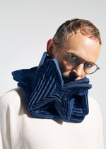 A person in glasses and a white top faces the camera, wearing the TYPE-O STOLE 002 by A-POC ABLE ISSEY MIYAKE—a large, sculptural dark blue pleated stole with origami folds. The background is plain and light-colored.