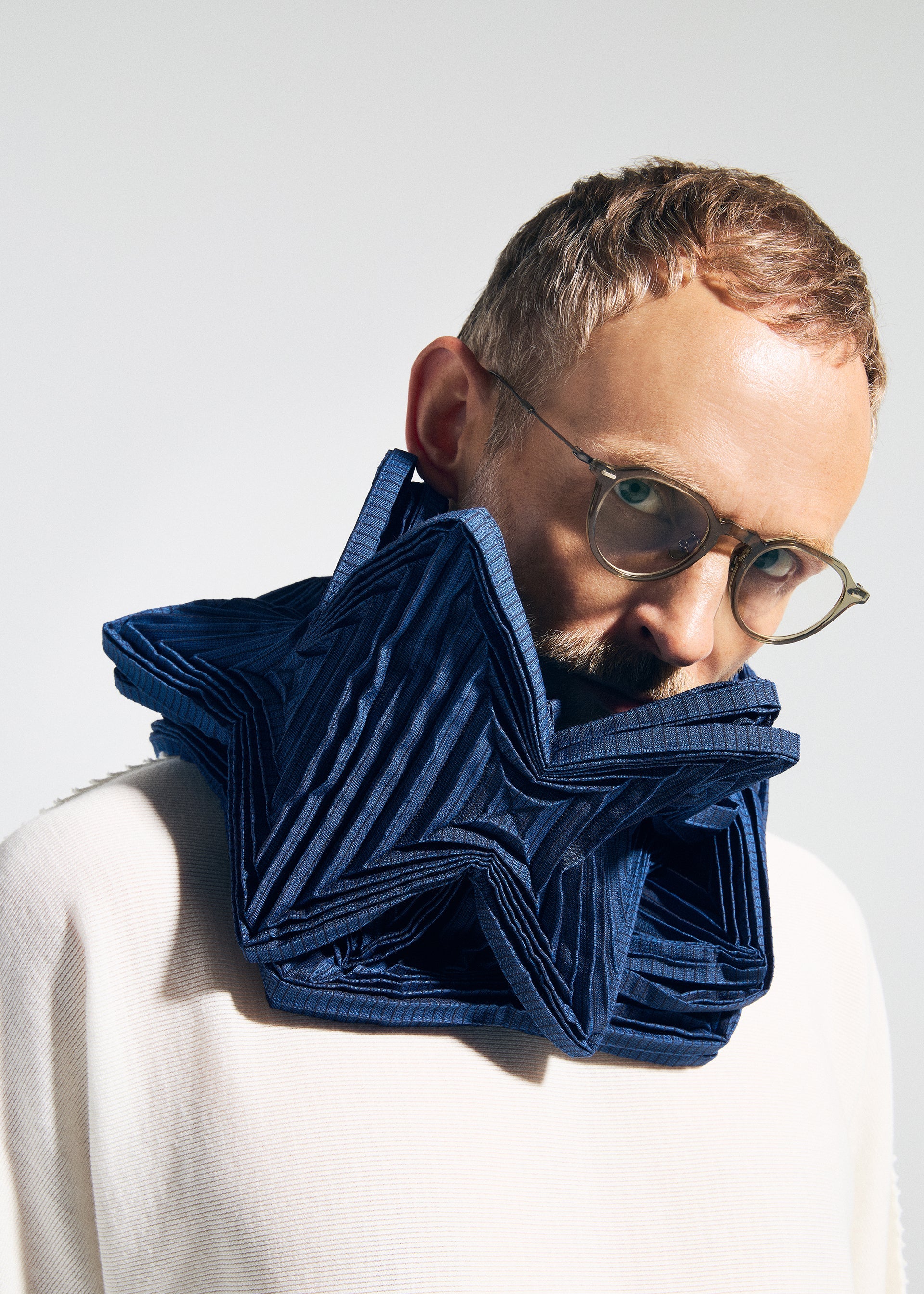 Zoomed view A person in glasses and a white top faces the camera, wearing the TYPE-O STOLE 002 by A-POC ABLE ISSEY MIYAKE—a large, sculptural dark blue pleated stole with origami folds. The background is plain and light-colored.