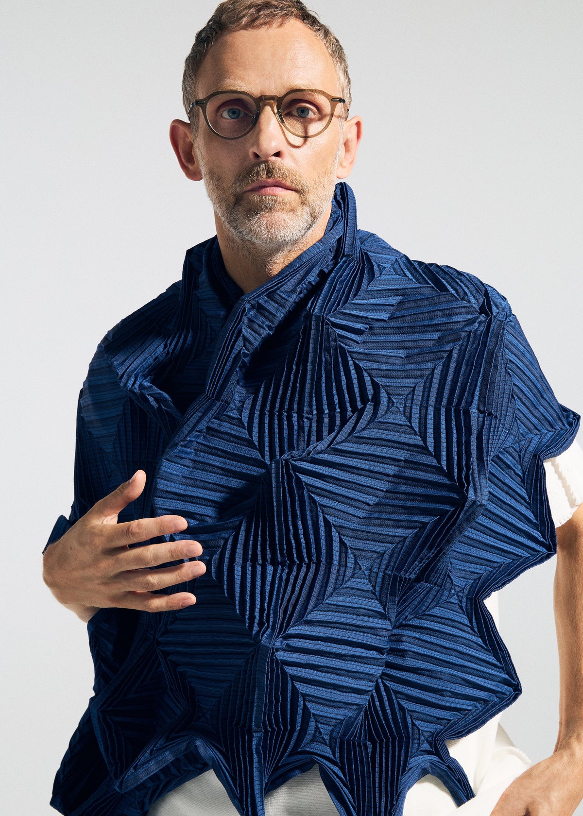 A man with short hair, glasses, and a beard wears the TYPE-O STOLE 002 by A-POC ABLE ISSEY MIYAKE—a sculptural blue stole with geometric patterns and origami folds—over a white shirt, standing against a neutral background. view 7 of 15