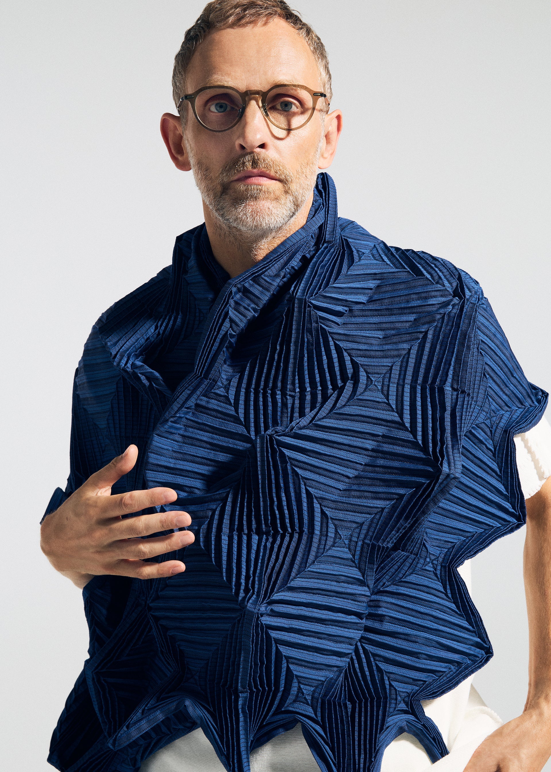 Zoomed view A man with short hair, glasses, and a beard wears the TYPE-O STOLE 002 by A-POC ABLE ISSEY MIYAKE—a sculptural blue stole with geometric patterns and origami folds—over a white shirt, standing against a neutral background.