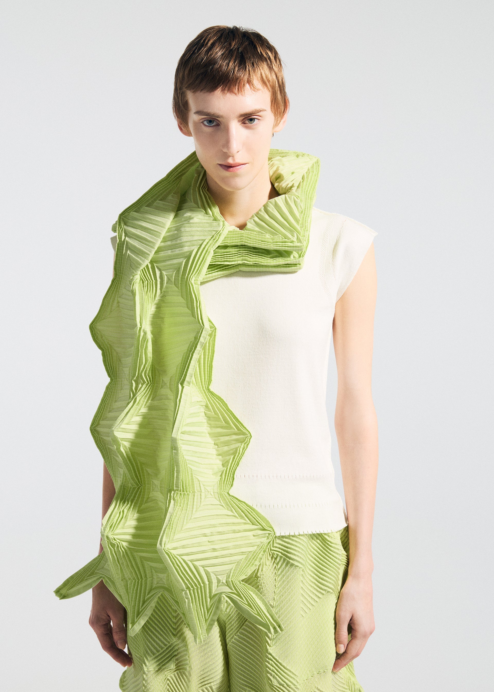 Zoomed view A person wears A-POC ABLE ISSEY MIYAKE's TYPE-O STOLE 002, a large green pleated stole draped origami-style over the shoulder, with a sleeveless white top and patterned light green pants against a plain light background.