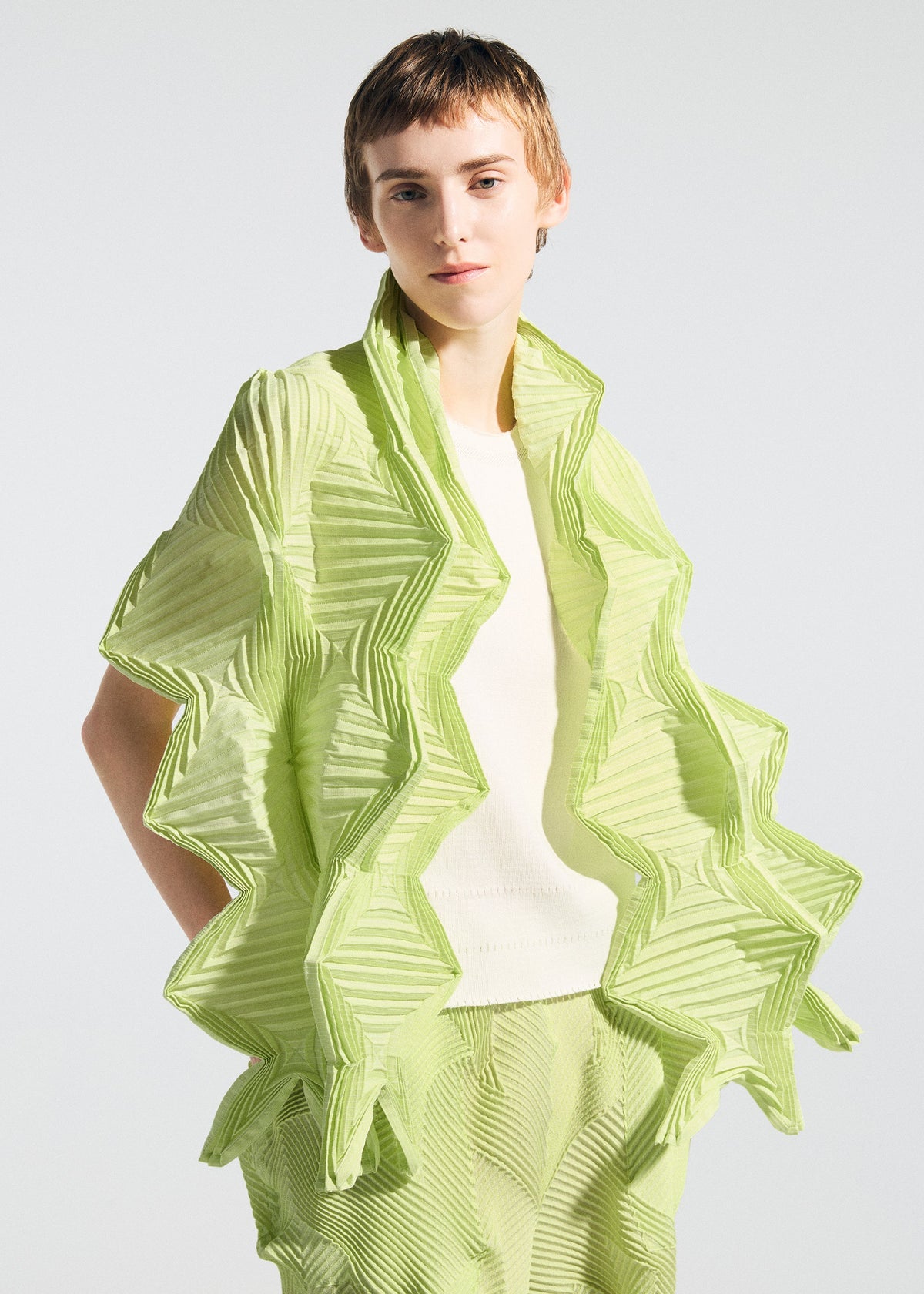 A person with short light brown hair wears the TYPE-O STOLE 002 by A-POC ABLE ISSEY MIYAKE, a textured origami fold lime green piece over a white top and matching green pants, standing against a plain light background. view 9 of 15
