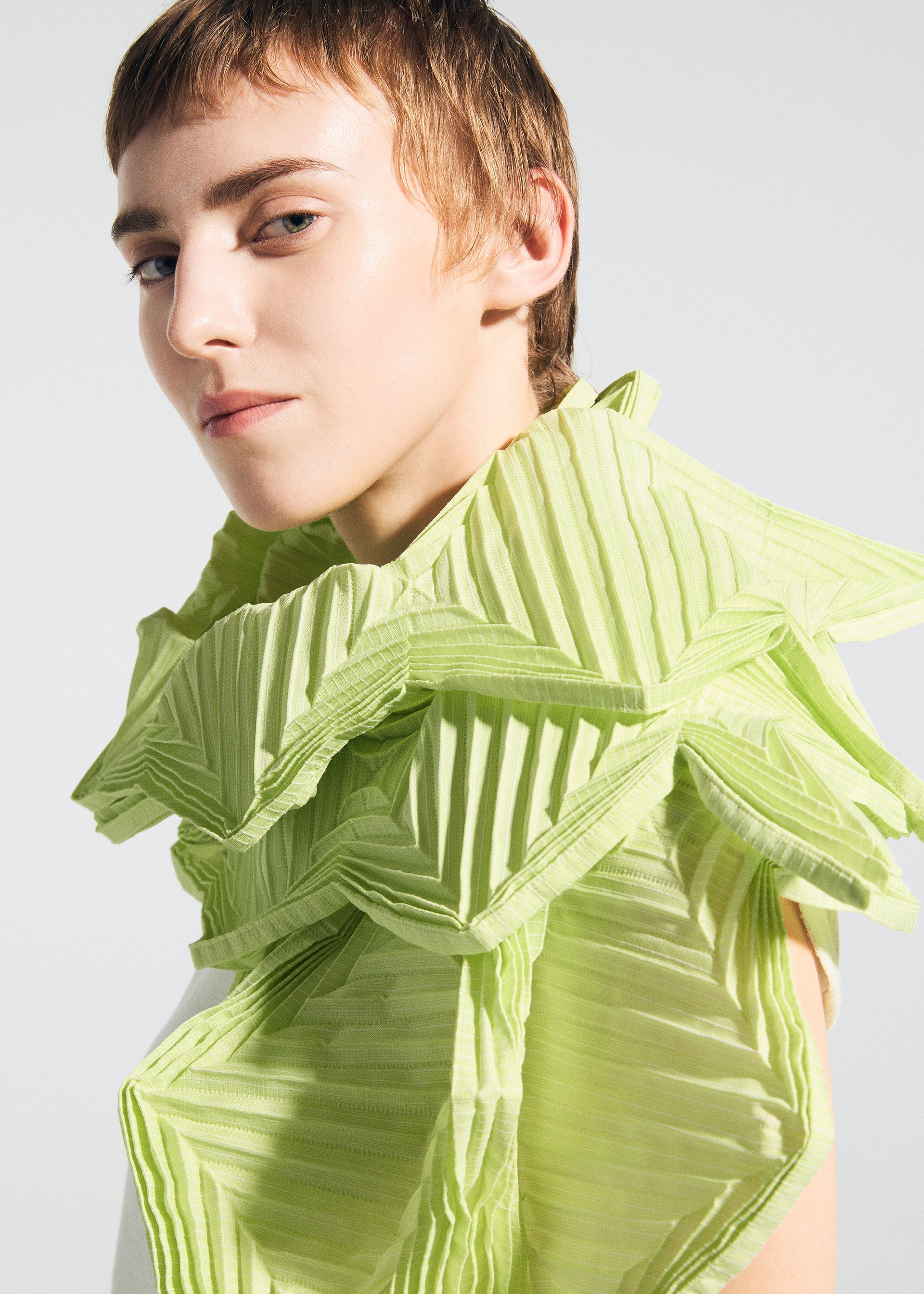 Zoomed view A person with short, light brown hair wears the TYPE-O STOLE 002 by A-POC ABLE ISSEY MIYAKE, a sculptural green piece with origami folds and layered ruffles, looking confidently at the camera against a plain light background.