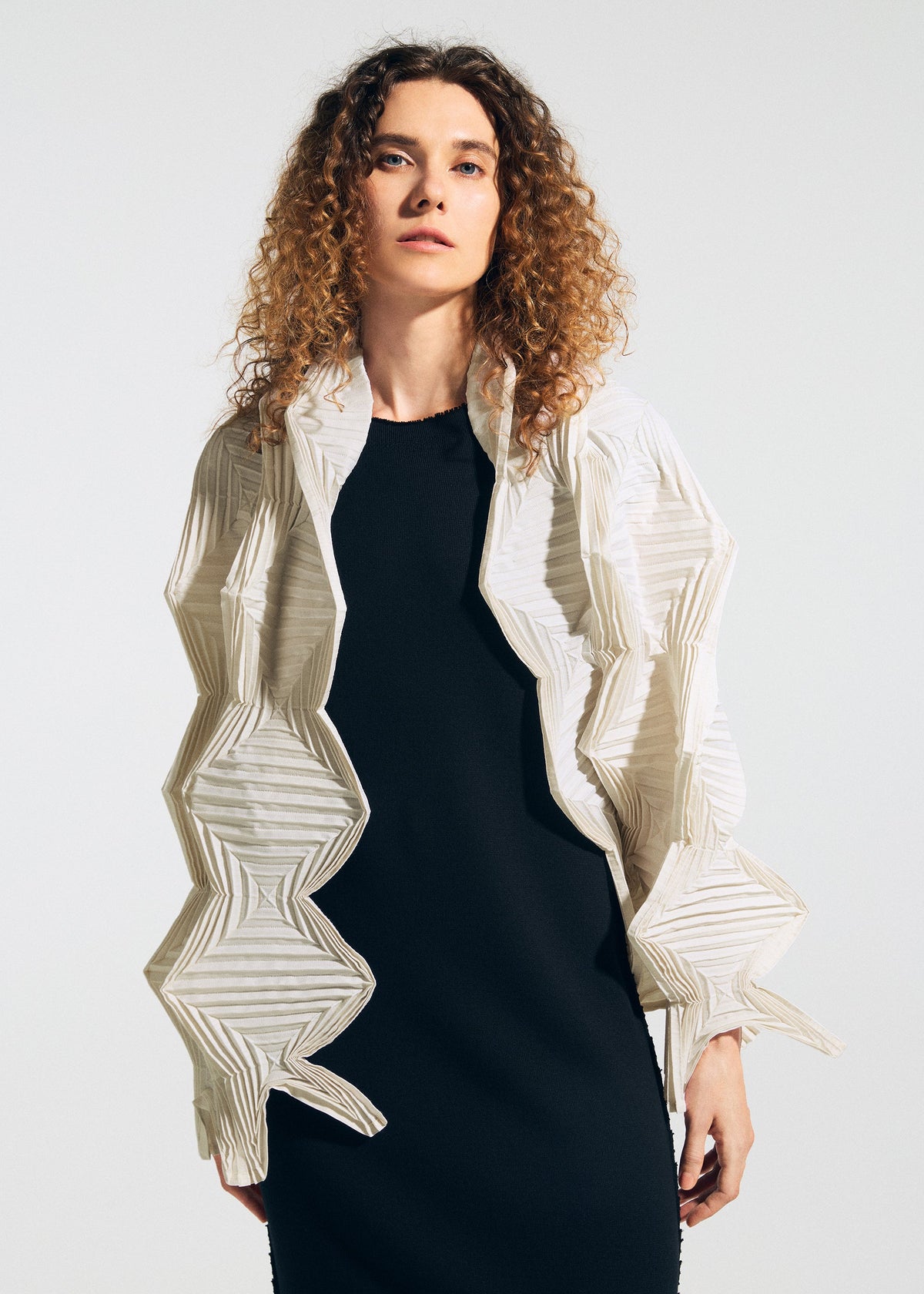 A person with curly hair wears a black dress and the TYPE-O STOLE 002 by A-POC ABLE ISSEY MIYAKE, a white geometric jacket with origami zigzag folds, standing against a plain light background. view 11 of 15