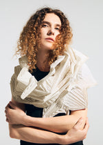 A person with curly brown hair poses with arms crossed, wearing the TYPE-O STOLE 002 in cream by A-POC ABLE ISSEY MIYAKE over a black top, set against a plain light background.