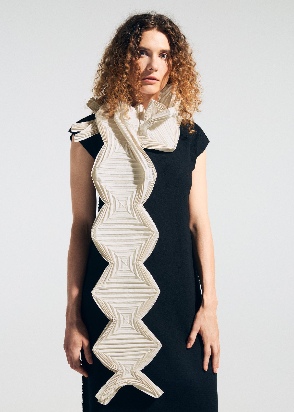 A woman with curly hair wears a sleeveless black dress and drapes the TYPE-O STOLE 002 by A-POC ABLE ISSEY MIYAKE—a large, geometric, origami-folded scarf in white and beige zigzag patterns—over her shoulder against a plain background. view 13 of 15