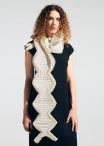 A woman with curly hair wears a sleeveless black dress and drapes the TYPE-O STOLE 002 by A-POC ABLE ISSEY MIYAKE—a large, geometric, origami-folded scarf in white and beige zigzag patterns—over her shoulder against a plain background.