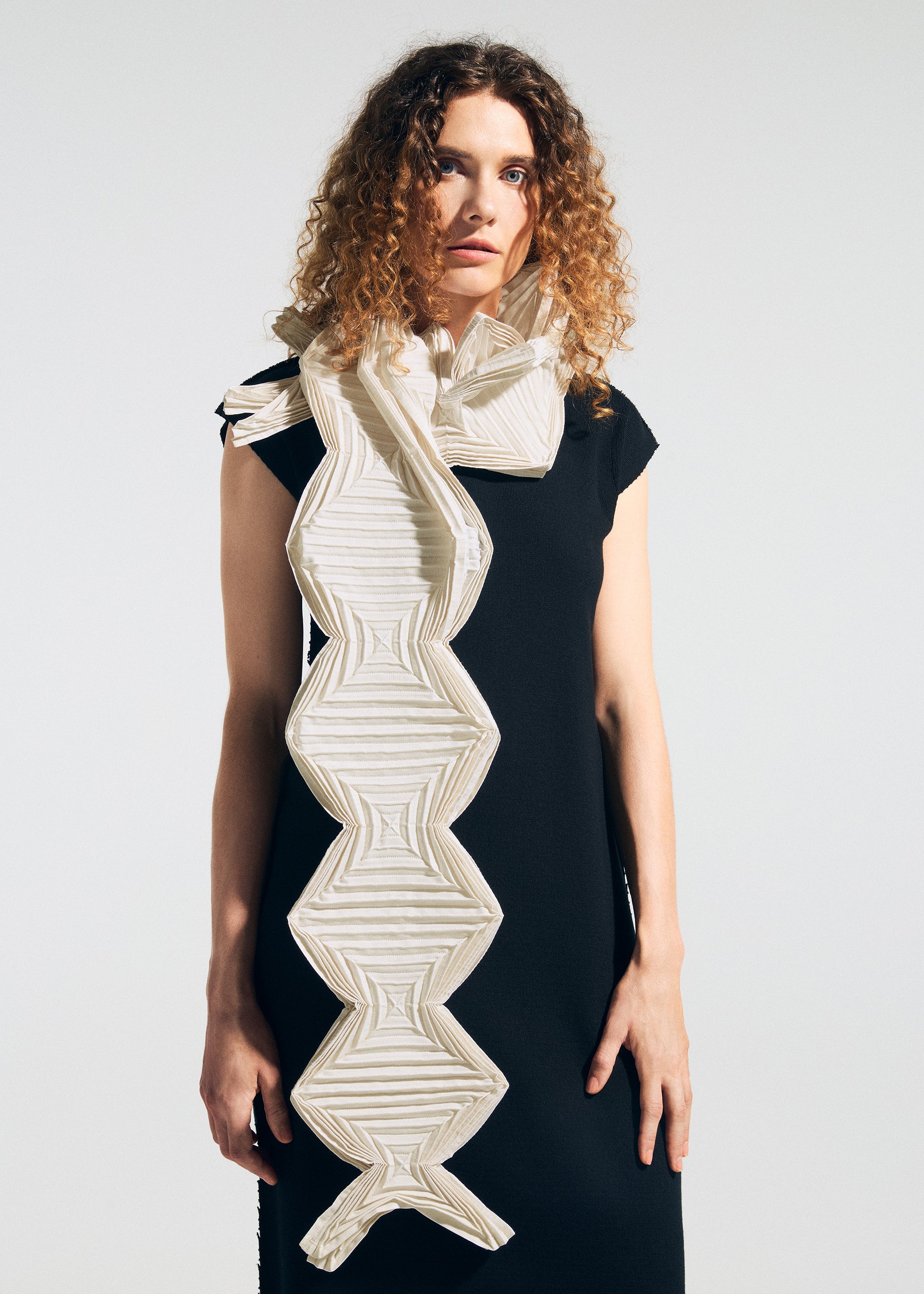 Zoomed view A woman with curly hair wears a sleeveless black dress and drapes the TYPE-O STOLE 002 by A-POC ABLE ISSEY MIYAKE—a large, geometric, origami-folded scarf in white and beige zigzag patterns—over her shoulder against a plain background.