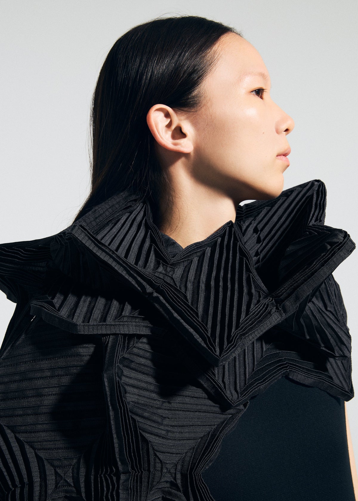 A woman with straight dark hair, shown in profile against a plain light background, wears a black outfit featuring the TYPE-O STOLE 002 by A-POC ABLE ISSEY MIYAKE, styled with dramatic pleated origami folds around her shoulders and neck. view 15 of 15