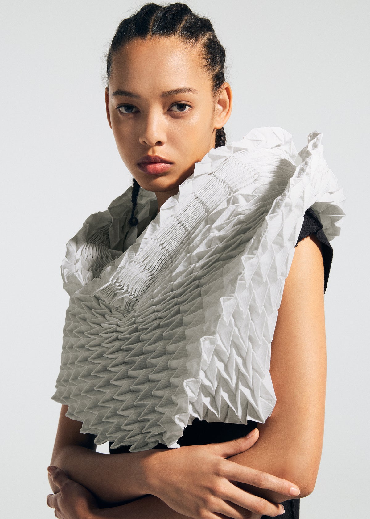 A person with braided hair wears a black top and TYPE-Ⅶ Kouzoshi project 002—a sculptural, geometric white collar crafted from folded material inspired by Tosa Washi paper by A-POC ABLE ISSEY MIYAKE—while standing against a plain background with a serious expression. view 12 of 15