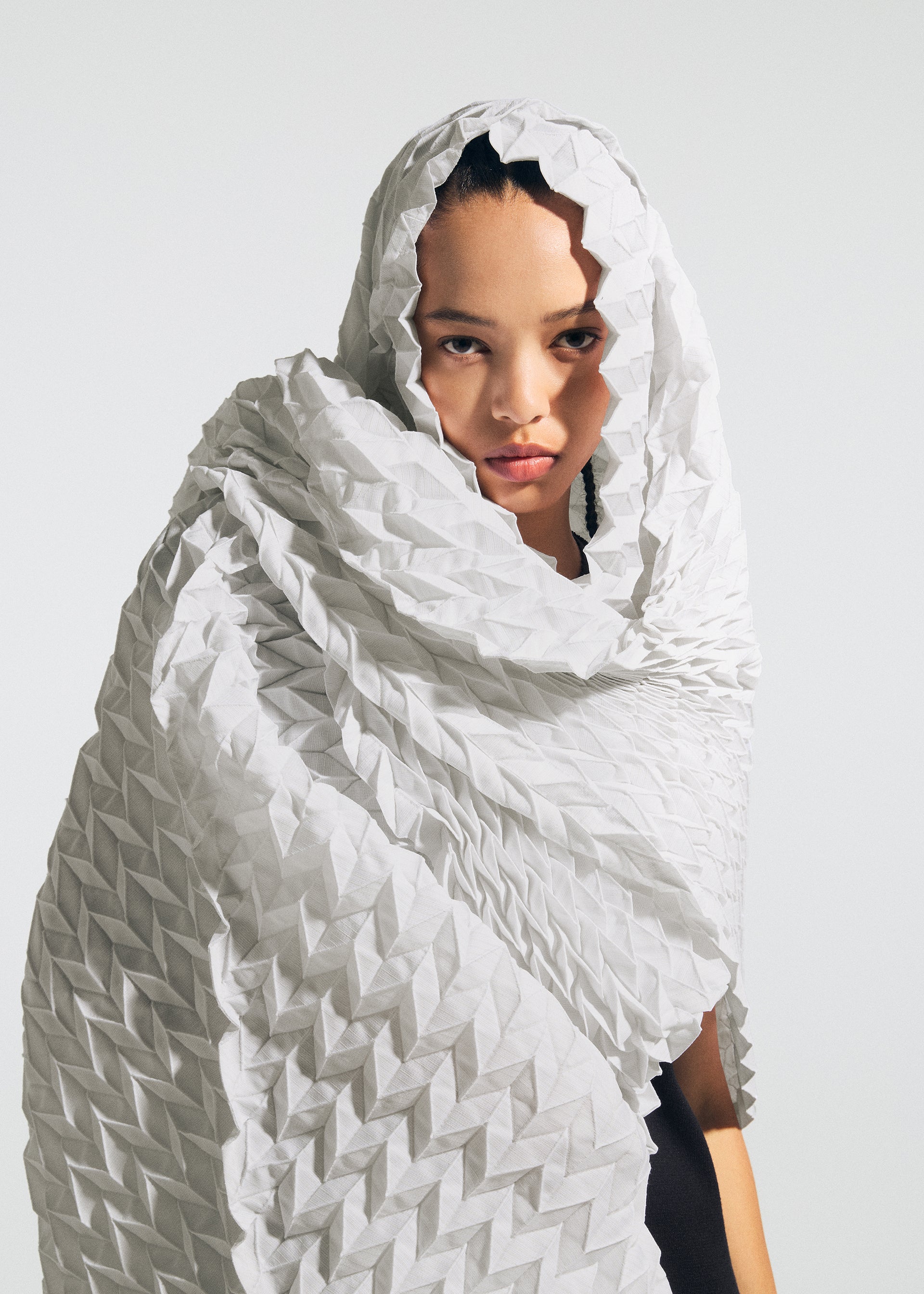 Zoomed view A person with a serious expression is wrapped in a textured, geometric white fabric reminiscent of Tosa Washi paper, elegantly enveloping their head and shoulders against a plain background—evoking the refined craftsmanship embodied in A-POC ABLE ISSEY MIYAKE’s TYPE-Ⅶ Kouzoshi project 002.