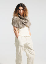 A person with curly hair stands with hands in pockets against a plain light background, wearing the TYPE-Ⅶ Kouzoshi project 002—an A-POC ABLE ISSEY MIYAKE stole featuring a textured geometric pattern, crafted from Tosa Washi paper—draped over a sleeveless white top and loose white pants.