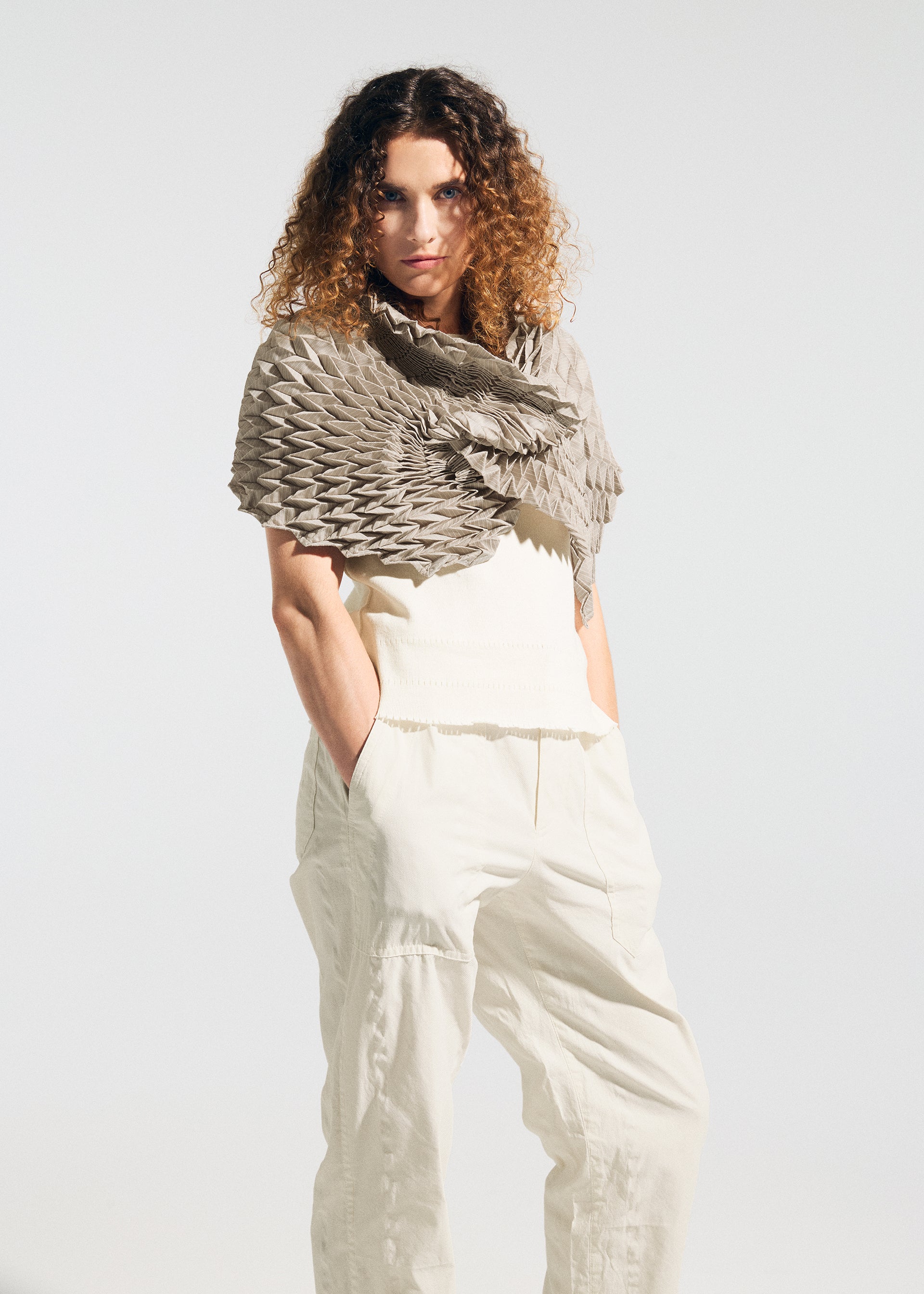 Zoomed view A person with curly hair stands with hands in pockets against a plain light background, wearing the TYPE-Ⅶ Kouzoshi project 002—an A-POC ABLE ISSEY MIYAKE stole featuring a textured geometric pattern, crafted from Tosa Washi paper—draped over a sleeveless white top and loose white pants.