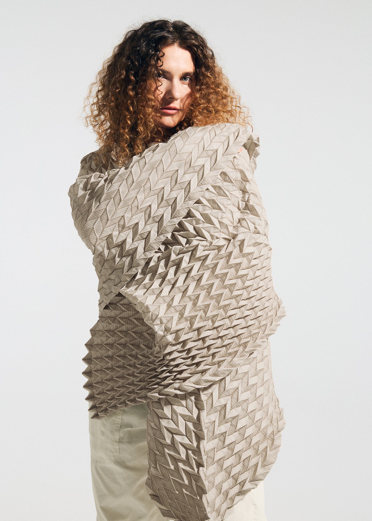 A person with curly hair, dressed in light-colored pants and draped in the TYPE-Ⅶ Kouzoshi project 002—a large, textured stole from A-POC ABLE ISSEY MIYAKE—featuring a bold geometric pattern, stands against a plain white background. view 6 of 15