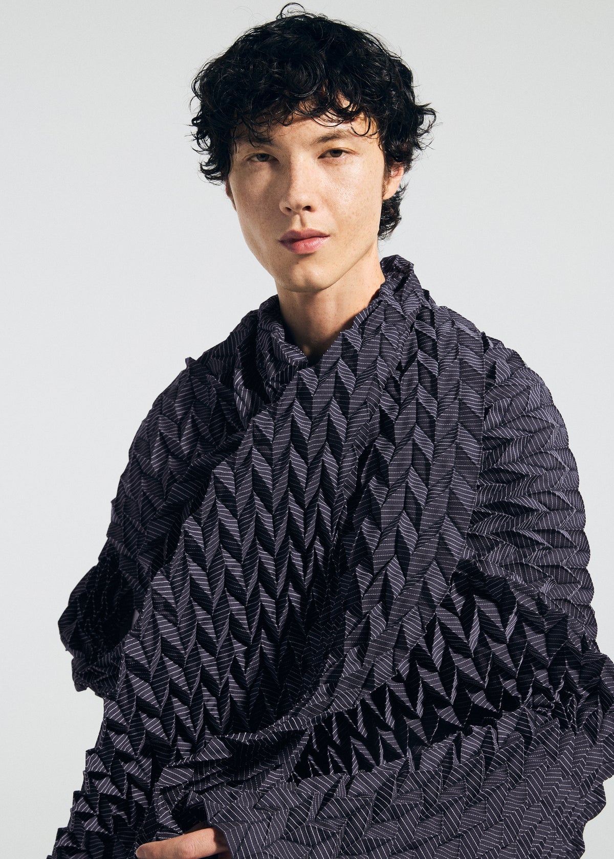 A person with short, curly black hair wears a textured, dark, patterned garment and a TYPE-Ⅶ Kouzoshi project 002 stole by A-POC ABLE ISSEY MIYAKE—crafted from Tosa Washi paper—and looks at the camera against a plain light background. view 7 of 15