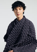 A person with short, curly black hair wears a textured, dark, patterned garment and a TYPE-Ⅶ Kouzoshi project 002 stole by A-POC ABLE ISSEY MIYAKE—crafted from Tosa Washi paper—and looks at the camera against a plain light background.