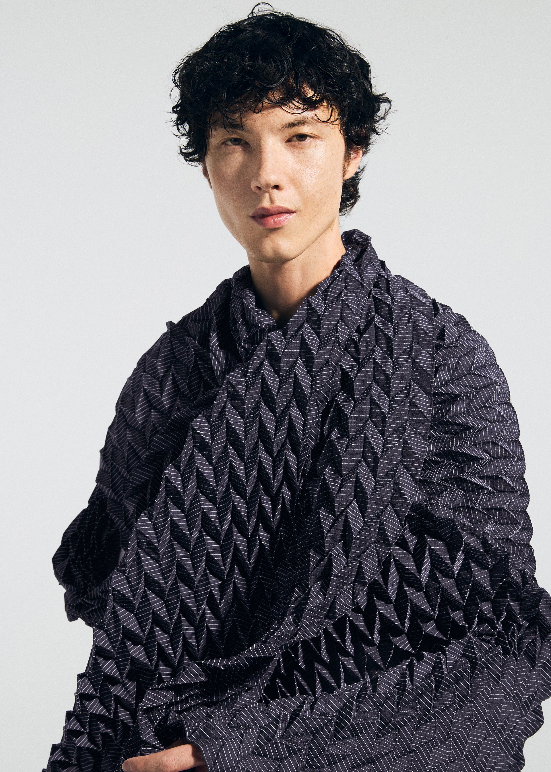 Zoomed view A person with short, curly black hair wears a textured, dark, patterned garment and a TYPE-Ⅶ Kouzoshi project 002 stole by A-POC ABLE ISSEY MIYAKE—crafted from Tosa Washi paper—and looks at the camera against a plain light background.