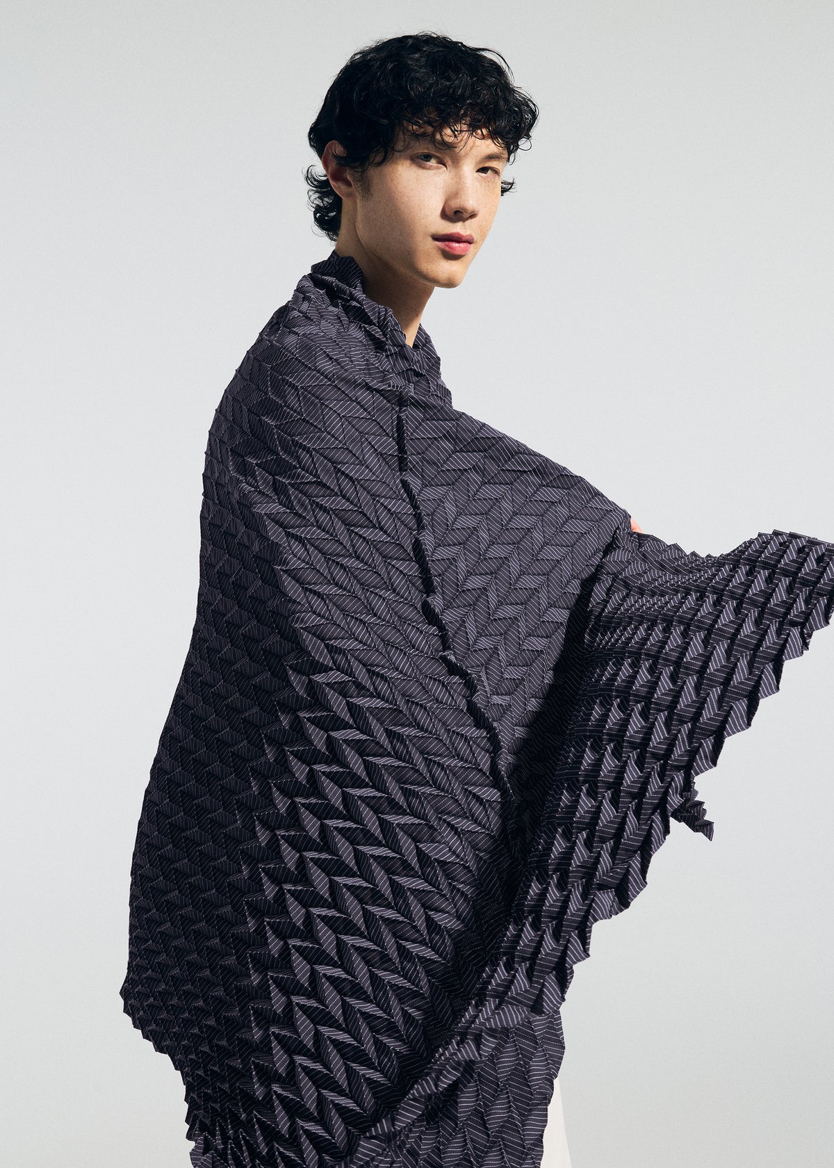 A person with curly dark hair wears the TYPE-Ⅶ Kouzoshi project 002 shawl by A-POC ABLE ISSEY MIYAKE, its textured geometric pattern draped elegantly around their shoulders. They stand against a light background, showcasing the refined craftsmanship of the piece. view 8 of 15