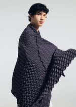 A person with curly dark hair wears the TYPE-Ⅶ Kouzoshi project 002 shawl by A-POC ABLE ISSEY MIYAKE, its textured geometric pattern draped elegantly around their shoulders. They stand against a light background, showcasing the refined craftsmanship of the piece.