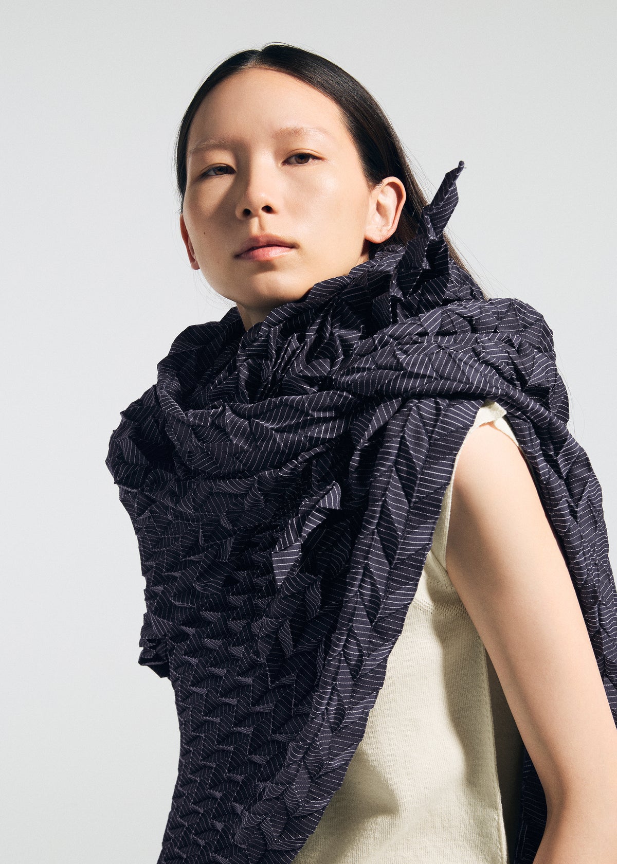 A person with long dark hair wears a textured, sculptural TYPE-Ⅶ Kouzoshi project 002 stole from A-POC ABLE ISSEY MIYAKE, draped over a sleeveless cream top, looking confidently at the camera against a plain light background. view 9 of 15