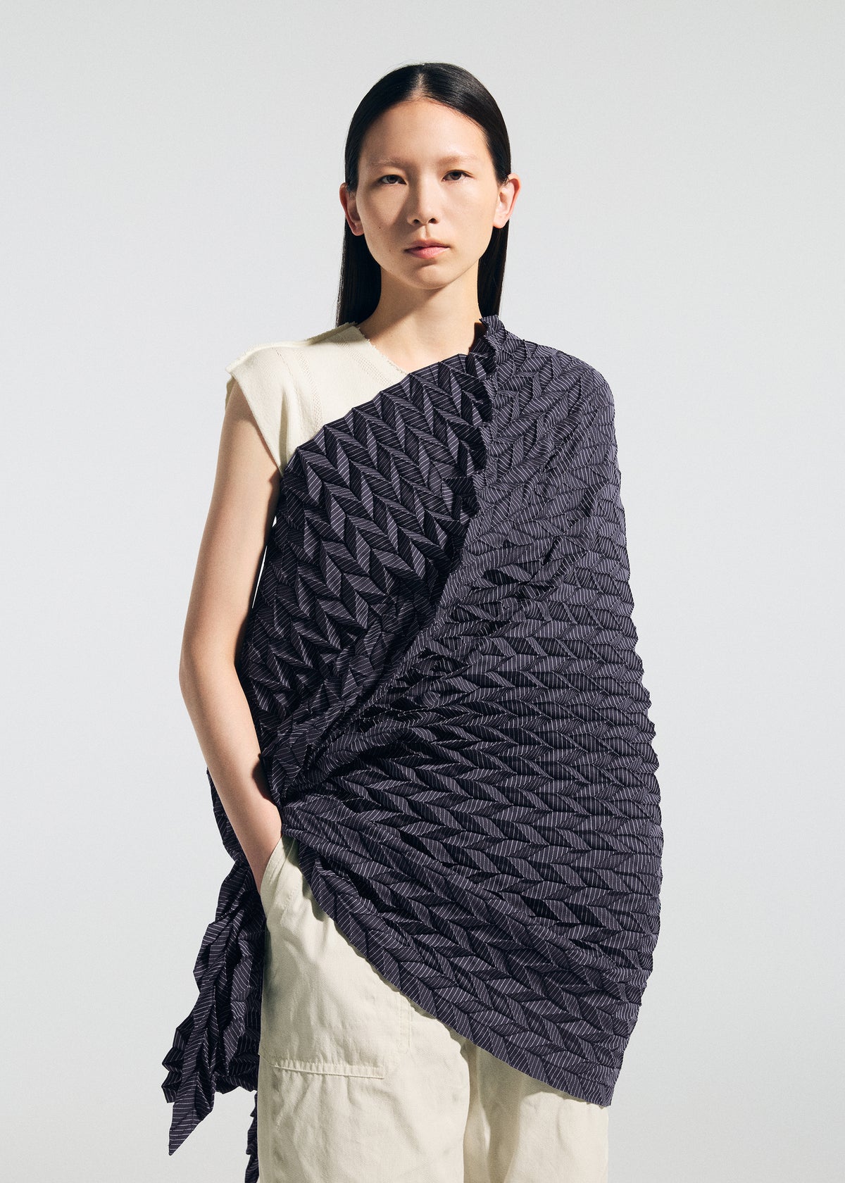 A person with long dark hair stands against a plain background, wearing the TYPE-Ⅶ Kouzoshi project 002—an intricately textured, dark geometric-patterned stole from A-POC ABLE ISSEY MIYAKE—draped over a light sleeveless top and light pants, hands in pockets. The look subtly highlights the brand’s innovative use of Tosa Washi paper in its collaborative design. view 10 of 15