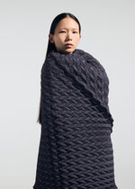 A person with long straight hair is wrapped in a textured, dark, geometric-patterned fabric reminiscent of the TYPE-Ⅶ Kouzoshi project 002 by A-POC ABLE ISSEY MIYAKE, standing against a plain light background and looking forward with a neutral expression.