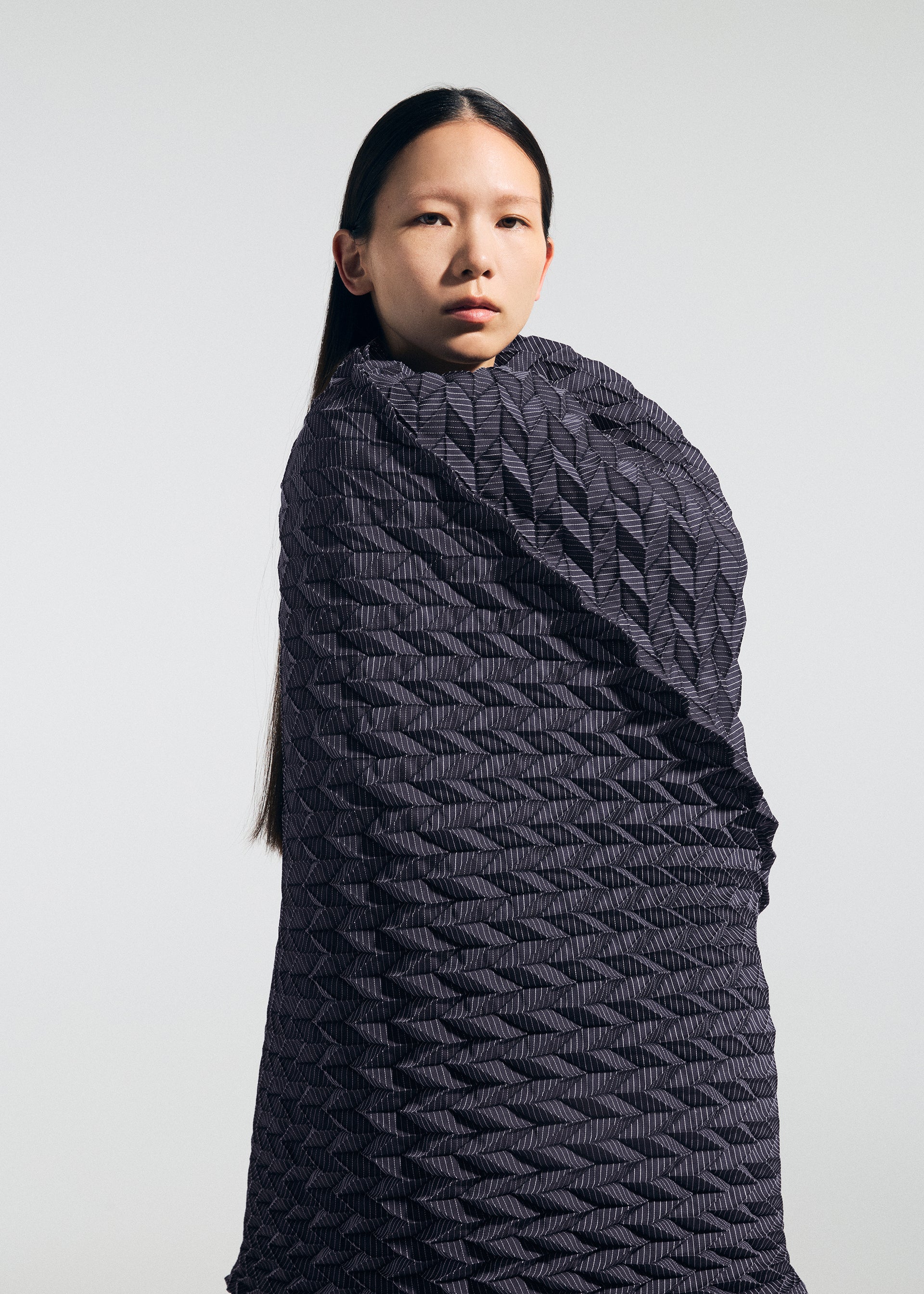 Zoomed view A person with long straight hair is wrapped in a textured, dark, geometric-patterned fabric reminiscent of the TYPE-Ⅶ Kouzoshi project 002 by A-POC ABLE ISSEY MIYAKE, standing against a plain light background and looking forward with a neutral expression.