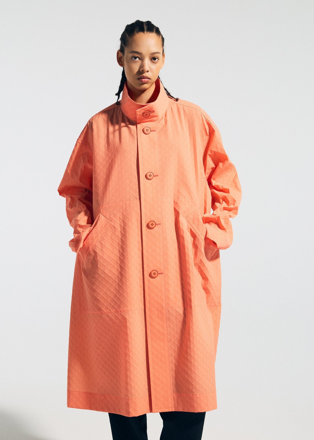 A person with braided hair wears the TYPE-T 001-1 coat by A-POC ABLE ISSEY MIYAKE, featuring a loose fit, unique textured fabric in coral with large pockets, standing against a plain light background. view 4 of 12