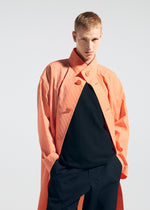 A person with short blond hair models the TYPE-T 001-1 coat by A-POC ABLE ISSEY MIYAKE in coral-orange, layered over a black shirt and dark pants, standing hands-in-pockets against a plain light backdrop.