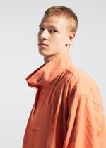 A young person with short blonde hair models the TYPE-T 001-1 coat by A-POC ABLE ISSEY MIYAKE, featuring a unique orange fabric and high collar, gazing confidently at the camera against a plain light background.