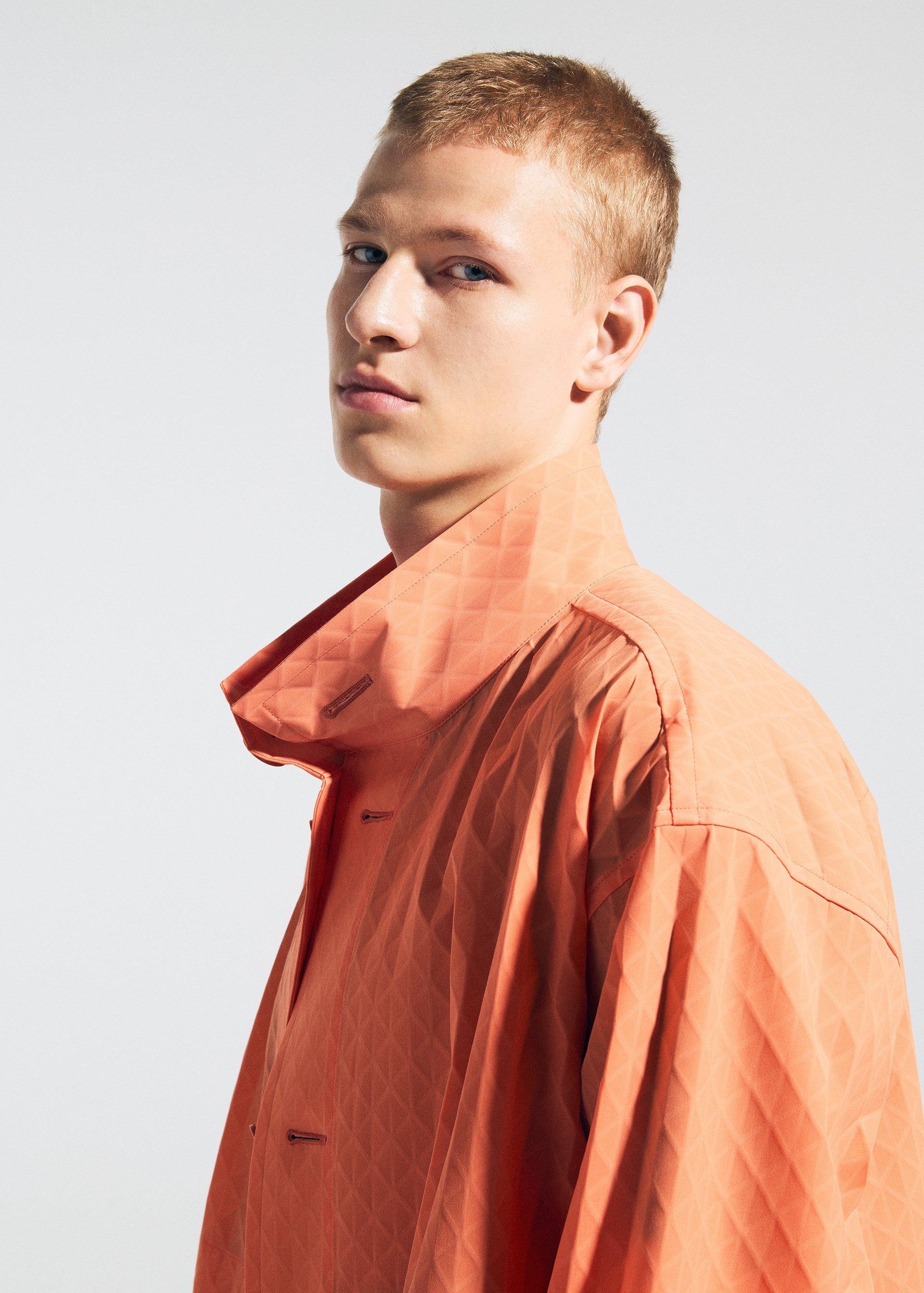 Zoomed view A young person with short blonde hair models the TYPE-T 001-1 coat by A-POC ABLE ISSEY MIYAKE, featuring a unique orange fabric and high collar, gazing confidently at the camera against a plain light background.