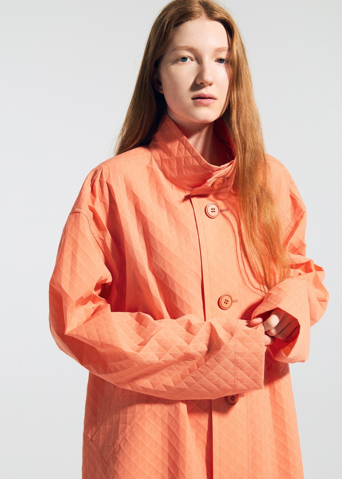 A person with long red hair models the TYPE-T 001-1 peach-colored coat by A-POC ABLE ISSEY MIYAKE, featuring large buttons and unique fabric, standing against a plain light background and looking slightly off-camera. view 7 of 12